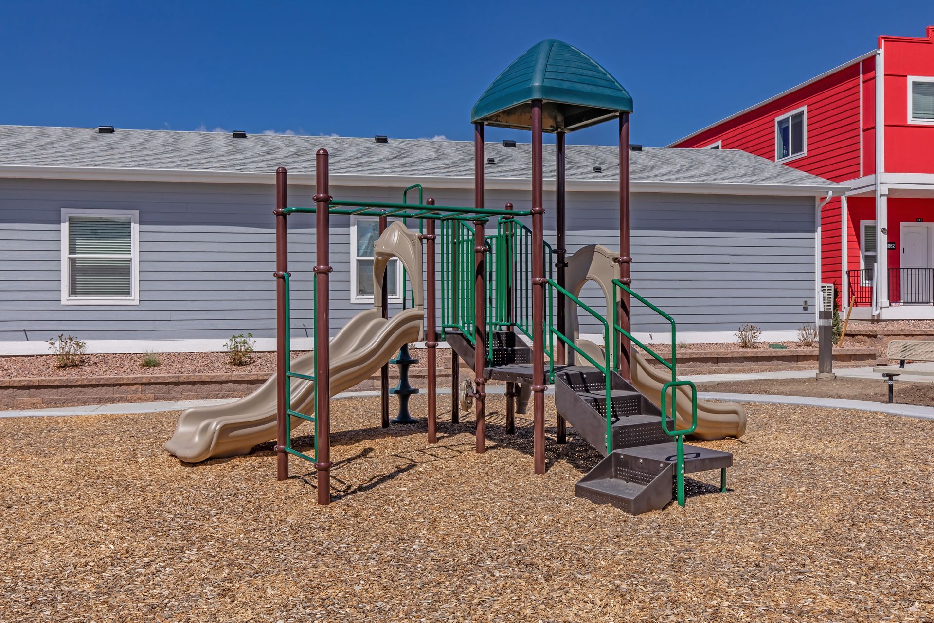 Children’s playground with slides and climbing structure on a gravel lot in front of a gray building