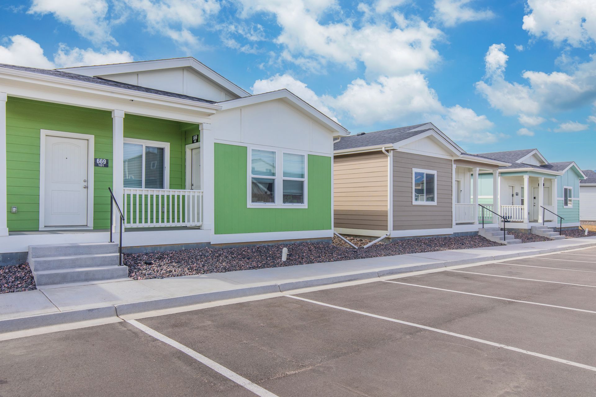 Row of colorful single-story houses with porches and empty parking spaces under a partly cloudy sky