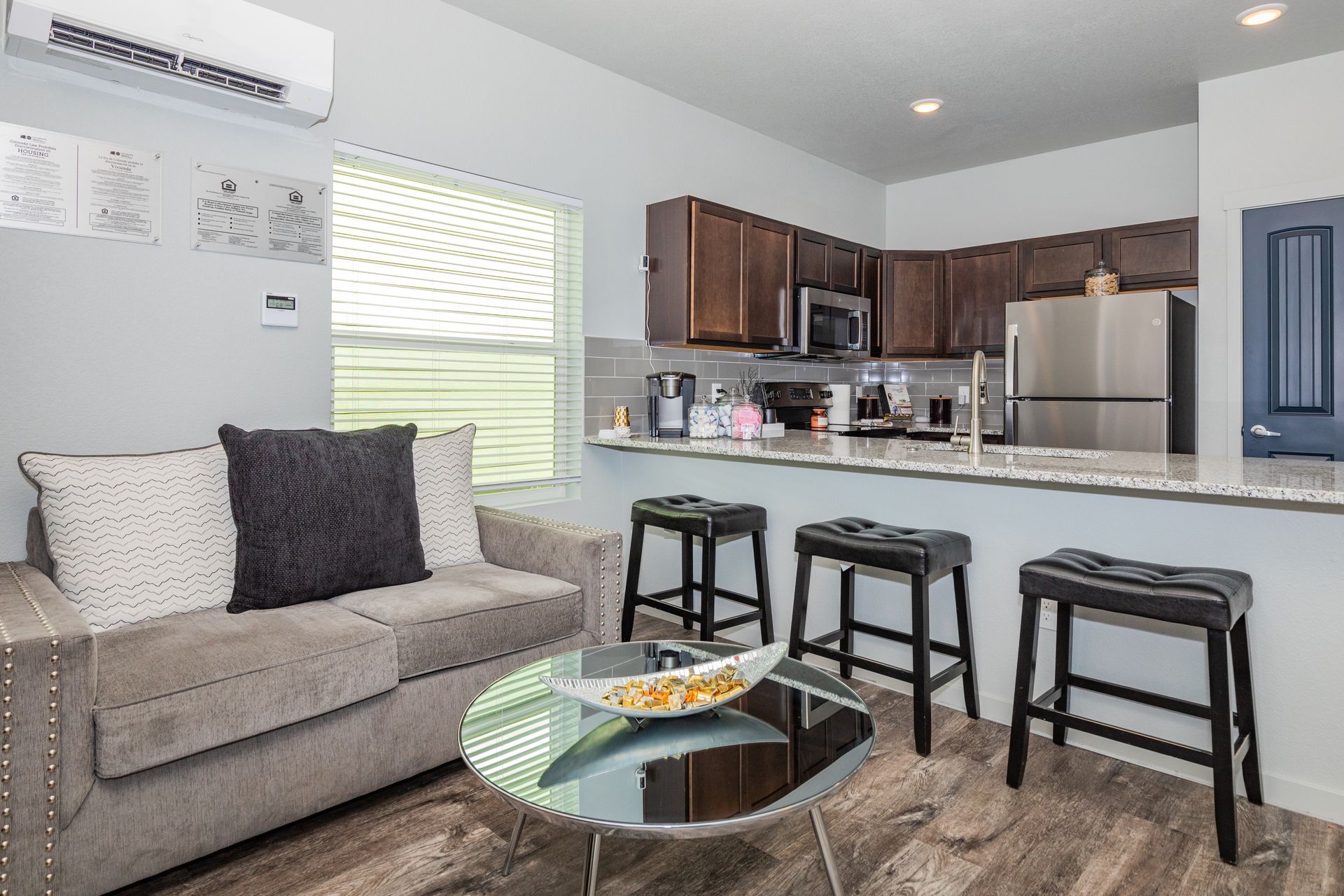 Small modern apartment living room and kitchen with gray sofa, glass coffee table, and bar stools.