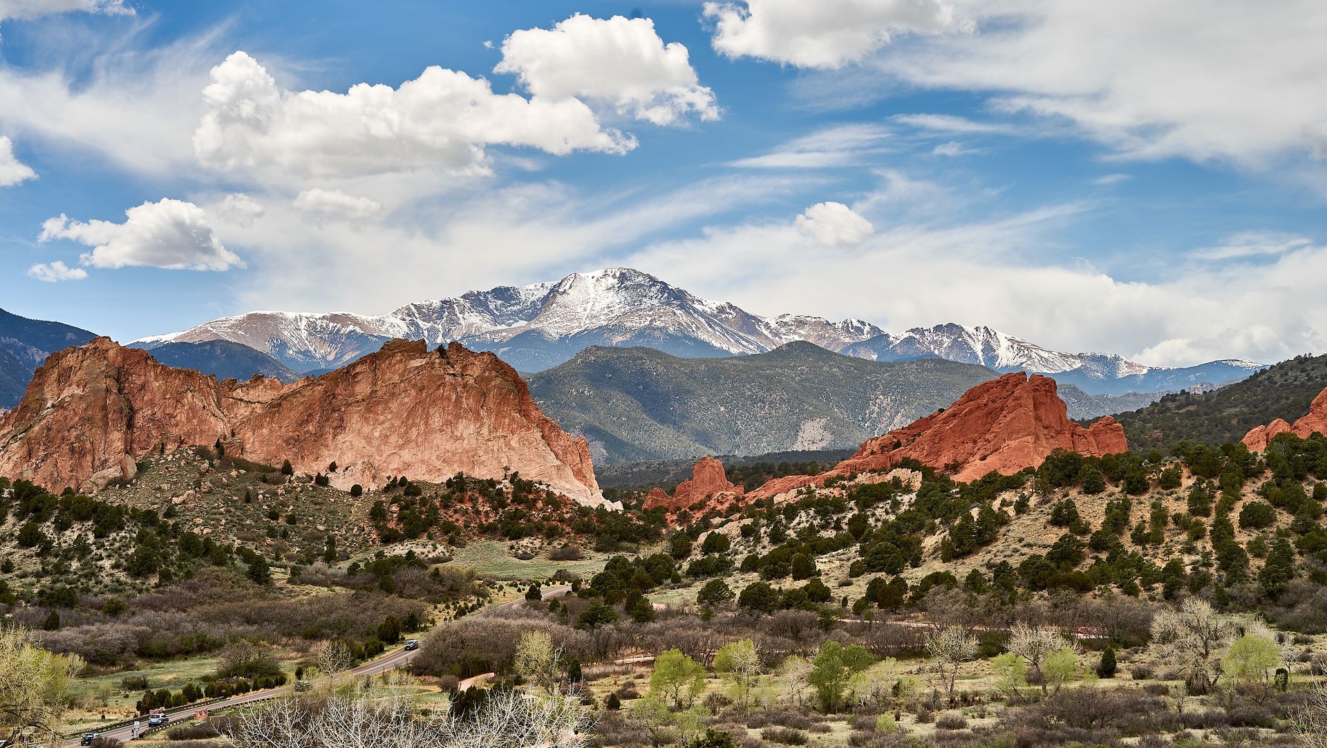 Red rock formations and snowy mountains under a partly cloudy sky above desert shrubs.