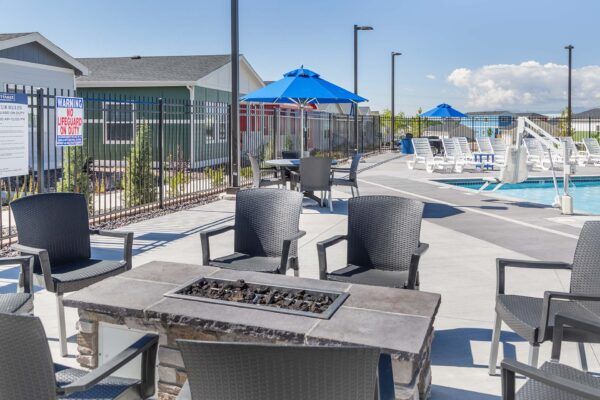 Outdoor pool patio with black chairs, fire pit, blue umbrellas, and lounge chairs by a fenced swimming pool