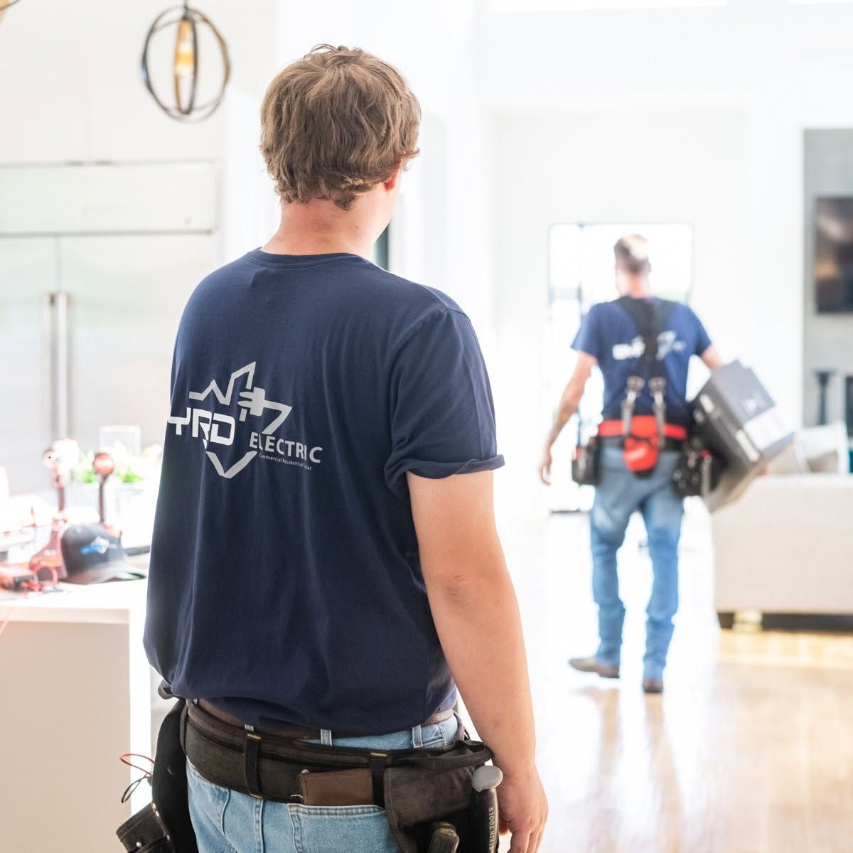Two workers in a bright room, one seen from behind in a navy shirt and tool belt.
