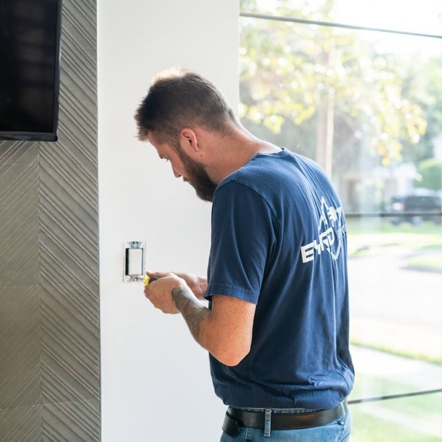 Technician in a blue shirt wiring a wall switch inside a bright room