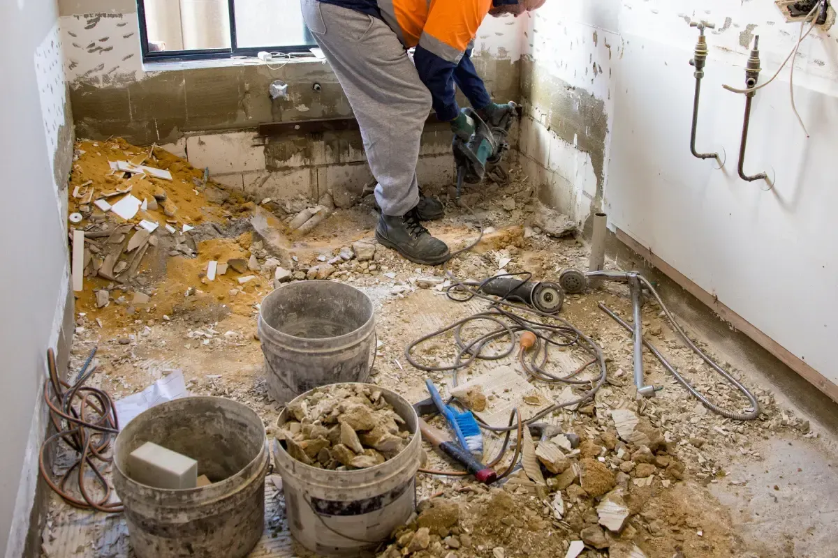 Person using a jackhammer inside a room undergoing renovation; debris, buckets, and tools are visible.