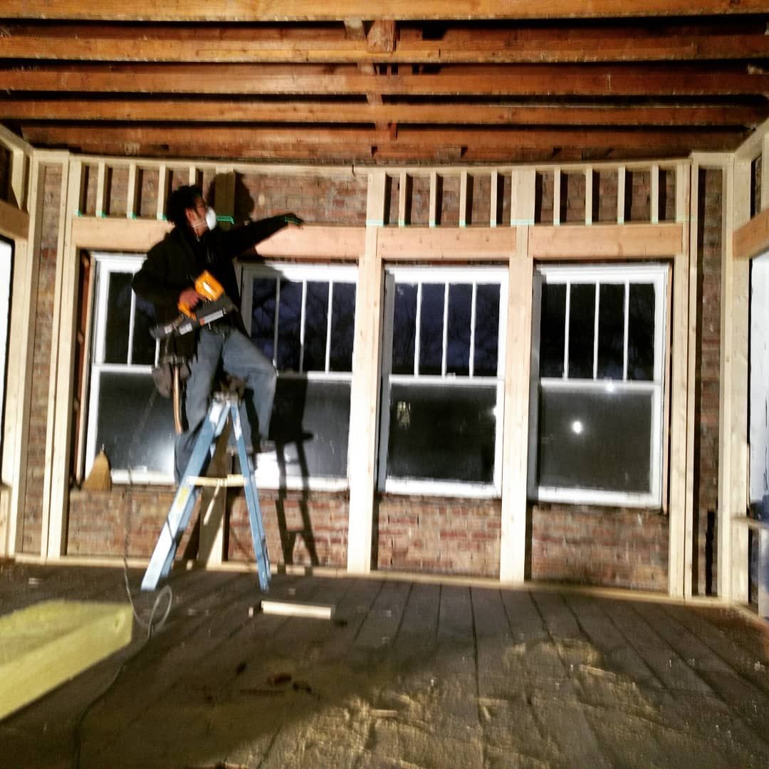Person on ladder in a room under construction, pointing with a tool towards window frames, exposed brick wall visible.
