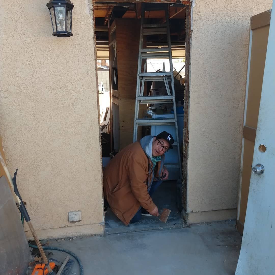Man working on doorway, wearing brown coat, with ladder inside; exterior setting.