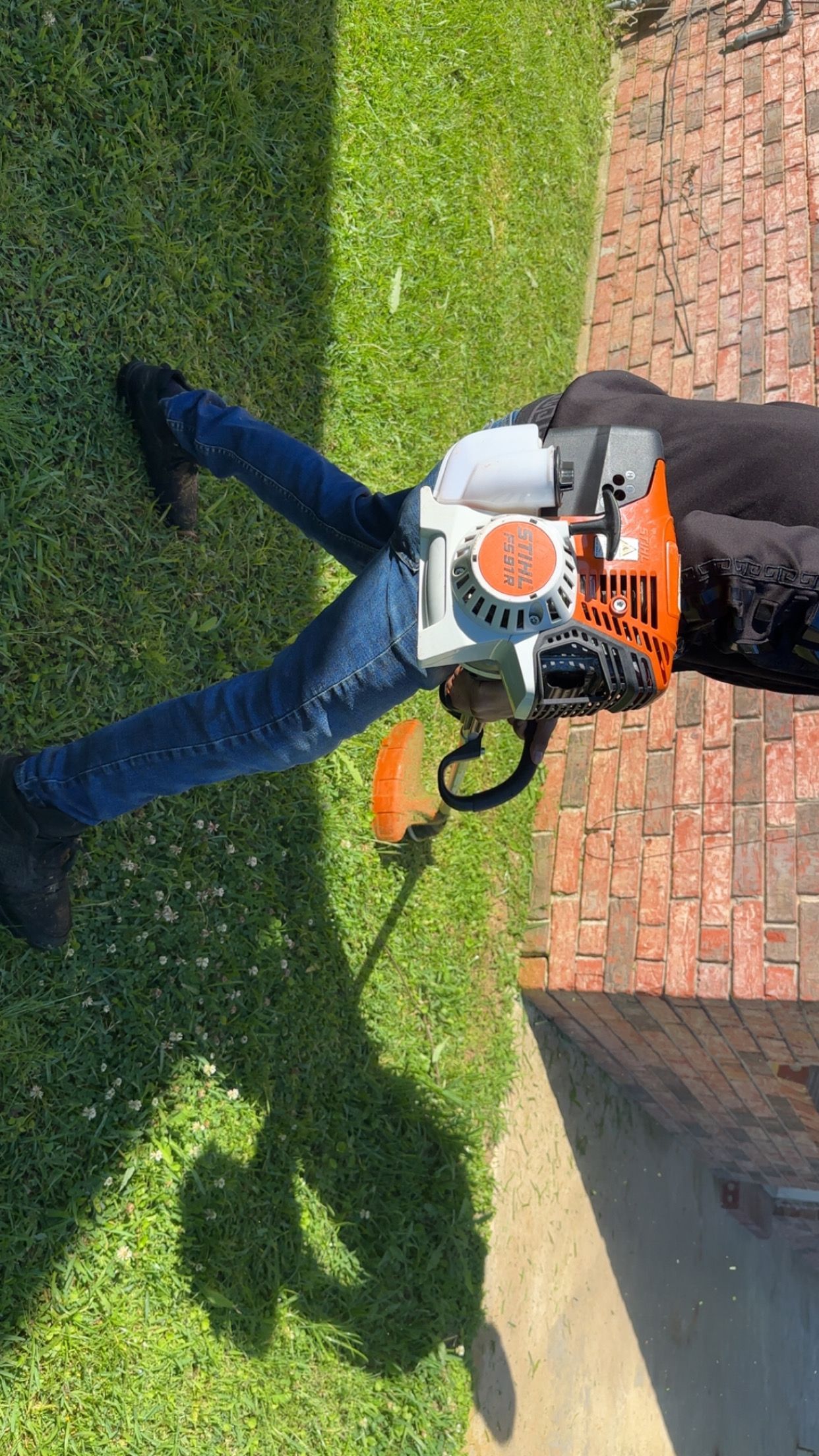 Person using an orange-and-white leaf blower on a lawn beside a brick wall.