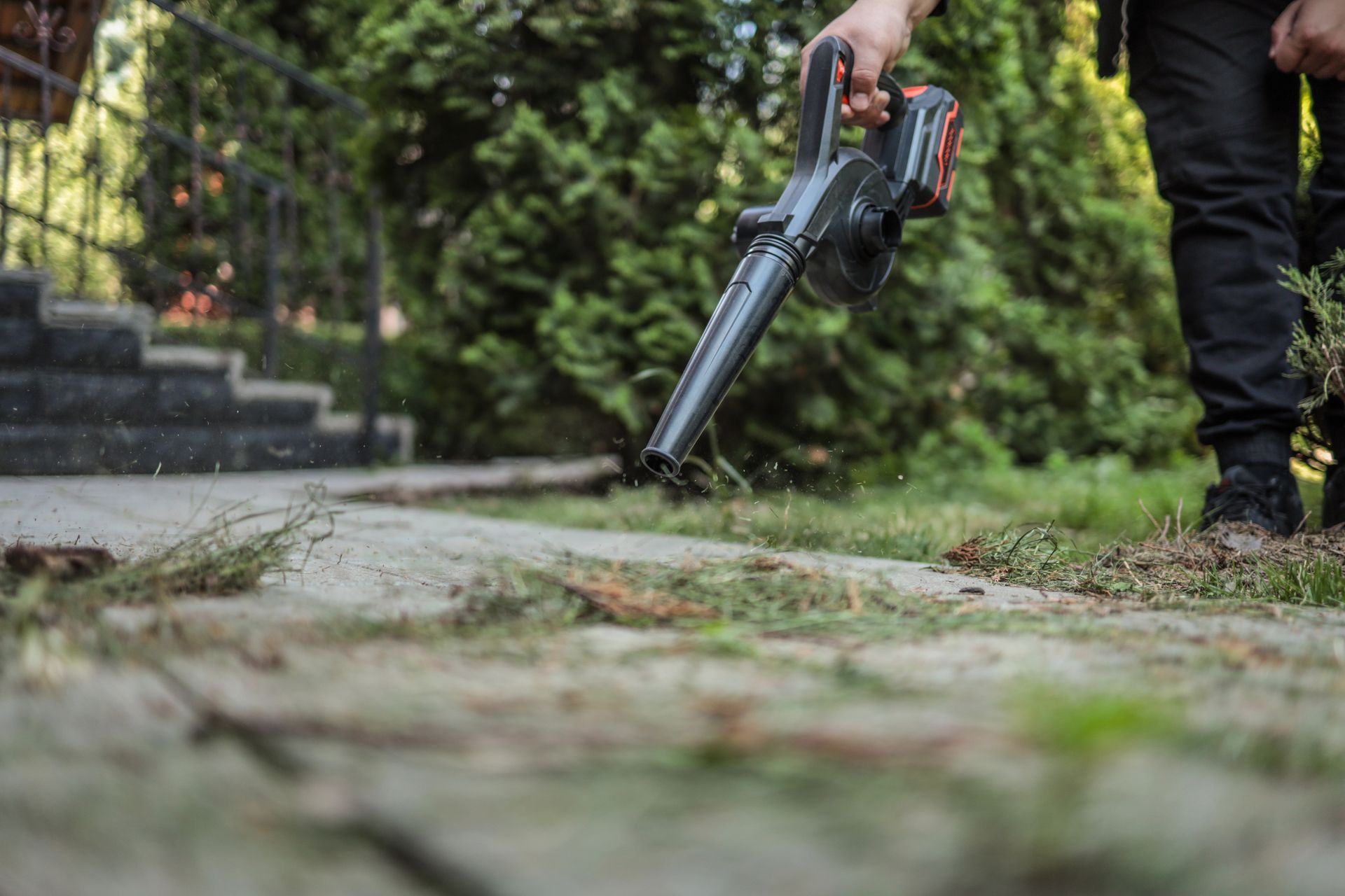 A person using a leaf blower to clear a garden path near stone steps and greenery