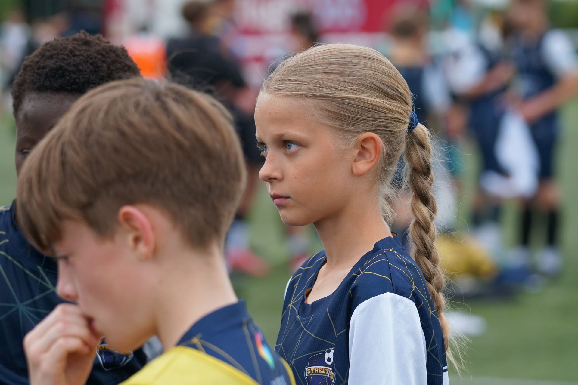 Two children in sports jerseys standing on a grassy field, one girl in profile with a braided ponytail.