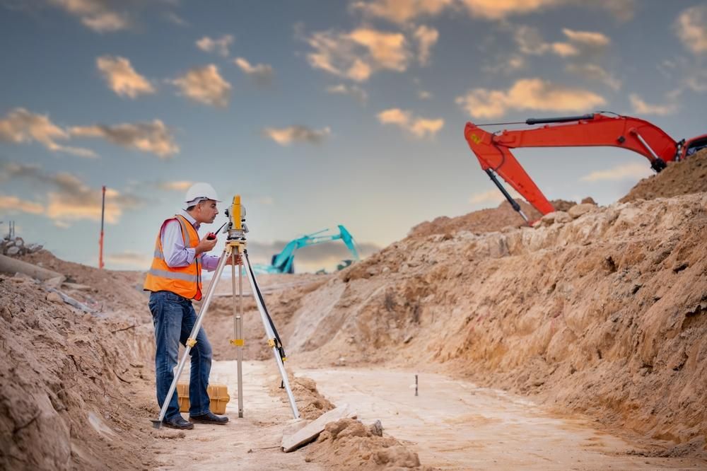 A Construction Worker Is Standing in The Dirt Using a Tripod — Duggan Mather Surveyors in Newcastle, NSW