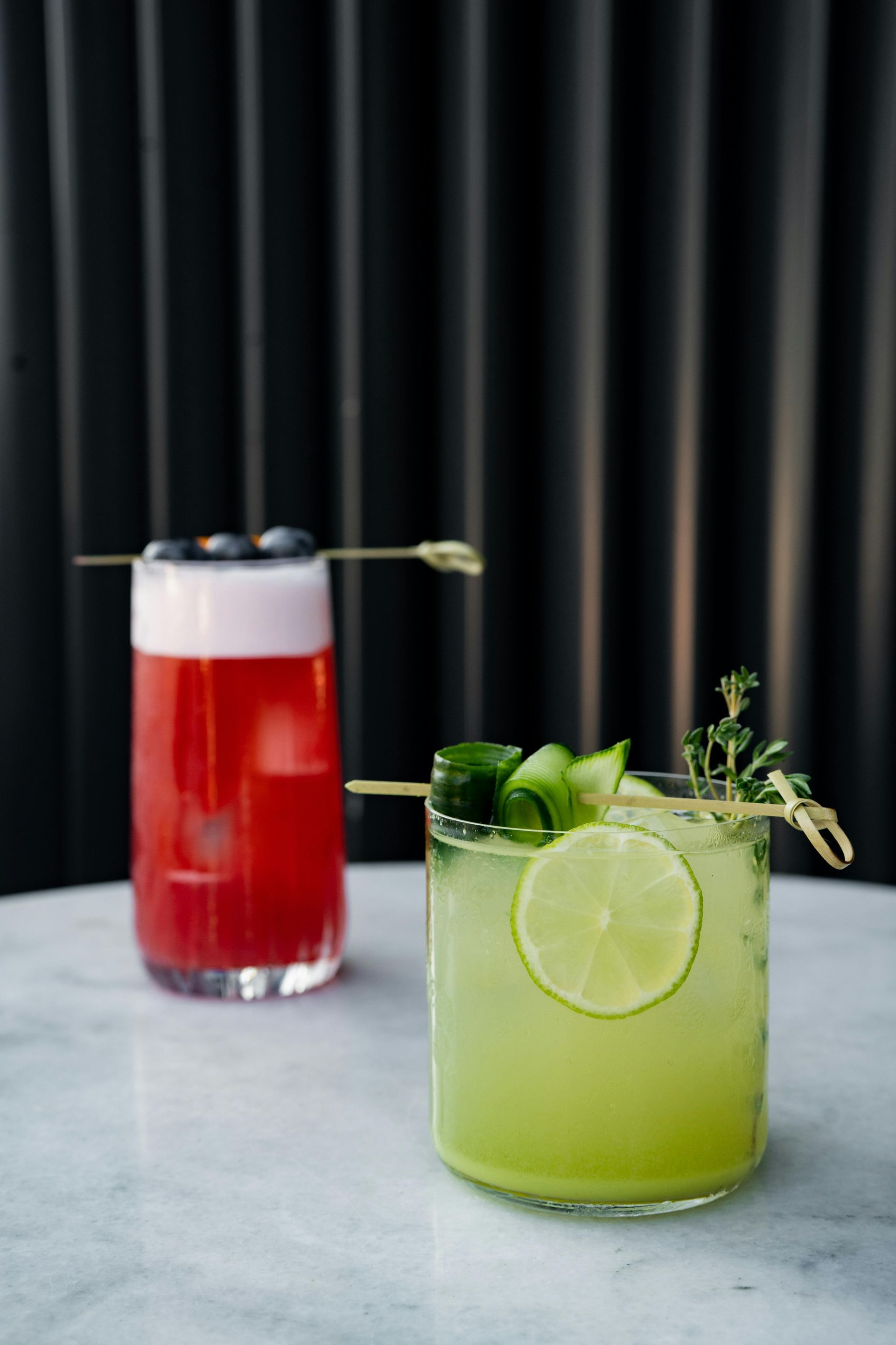 Two cocktails: one red with blueberries, one green with lime, on a white table.