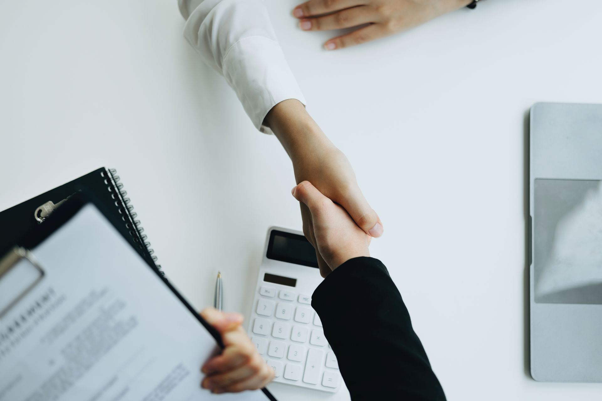 Two people shaking hands over a white table with a contract, calculator, and laptop.