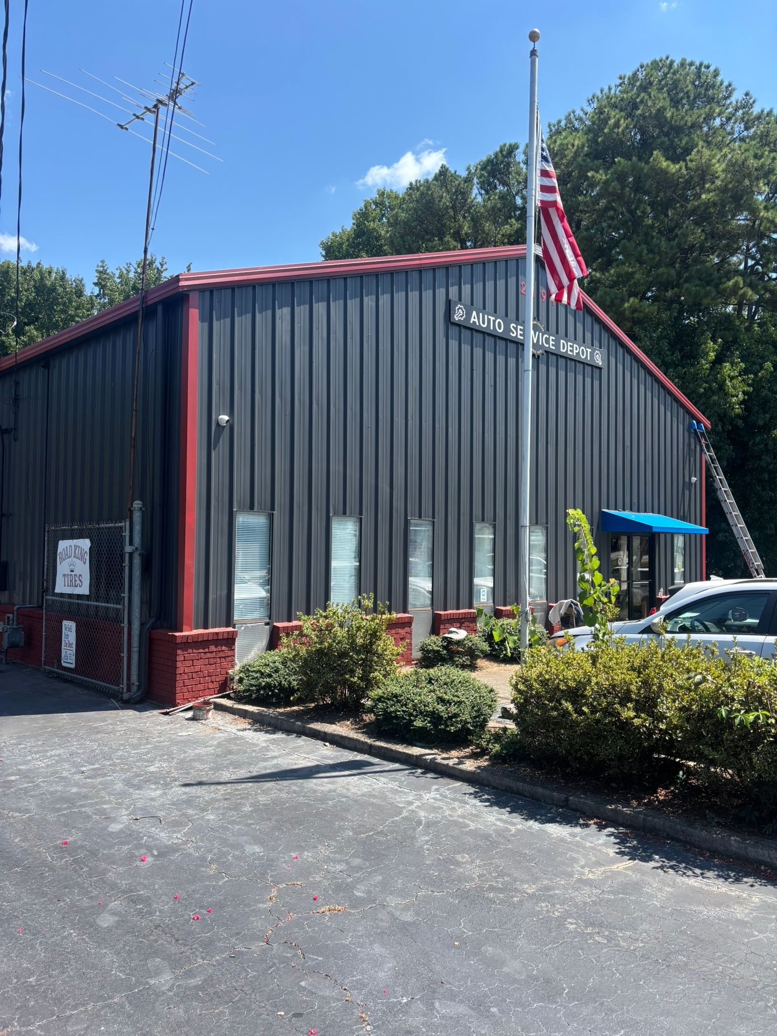 Small black building with red trim, American flag, and parked car by a hedge in sunlight | Auto Service Depot
