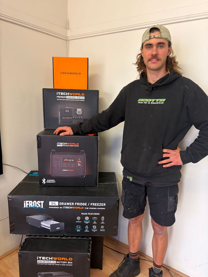 Man Standing Beside Stacked Product Boxes in a Corner of a Room, Wearing a Black Hoodie and Shorts — Scotts Auto Air & Electrical In Wollongong, NSW