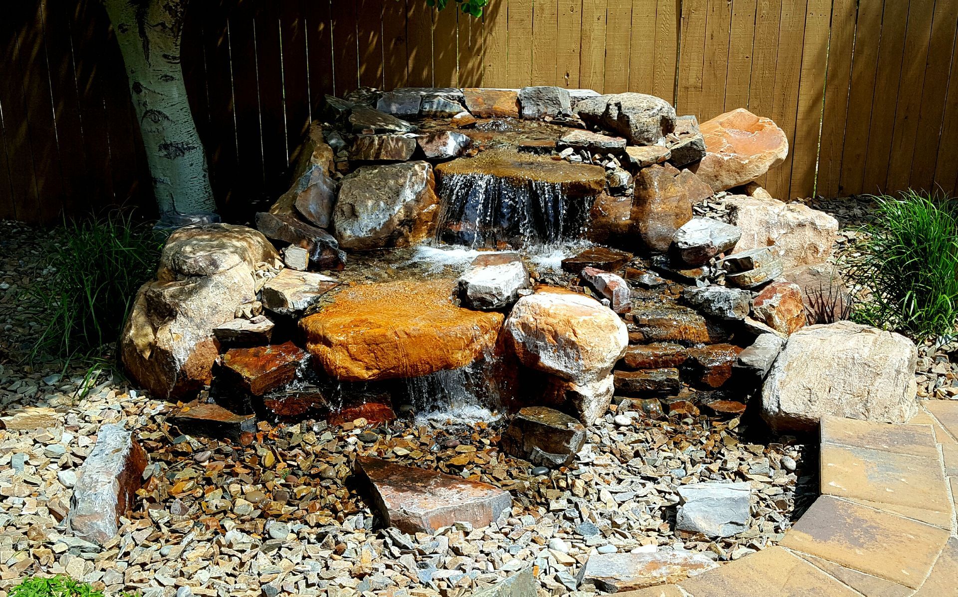 A small waterfall is surrounded by rocks and mulch in front of a house.