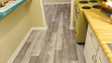 Gray wood-look flooring in a kitchen and hallway, yellow cabinets and a stove are in the kitchen.