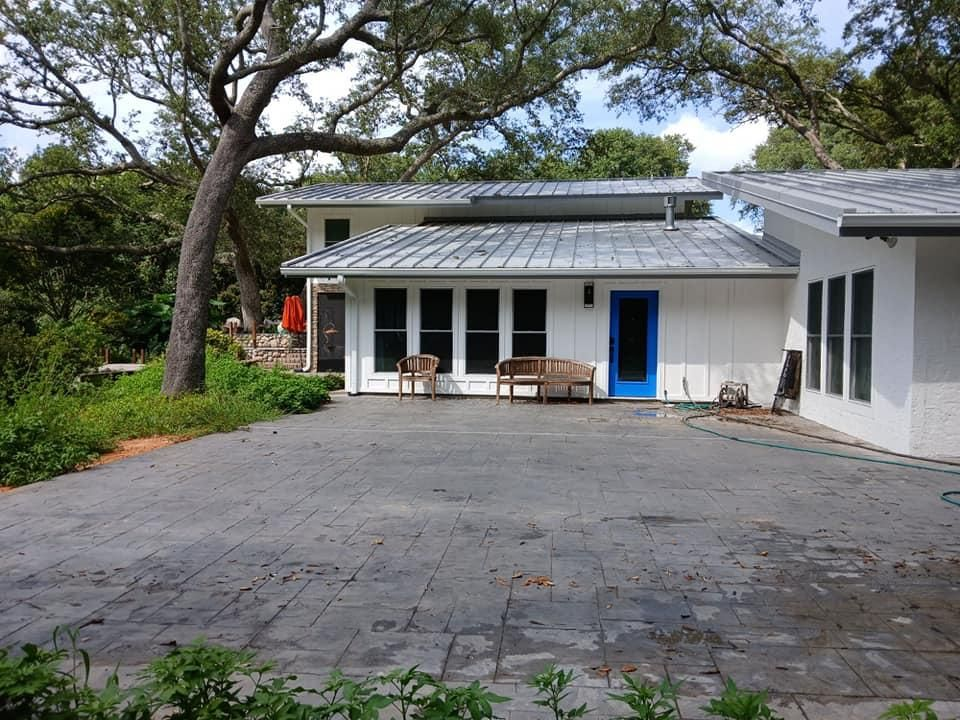 White house with blue door, grey roof, on a grey paved patio. Lush trees in the background.