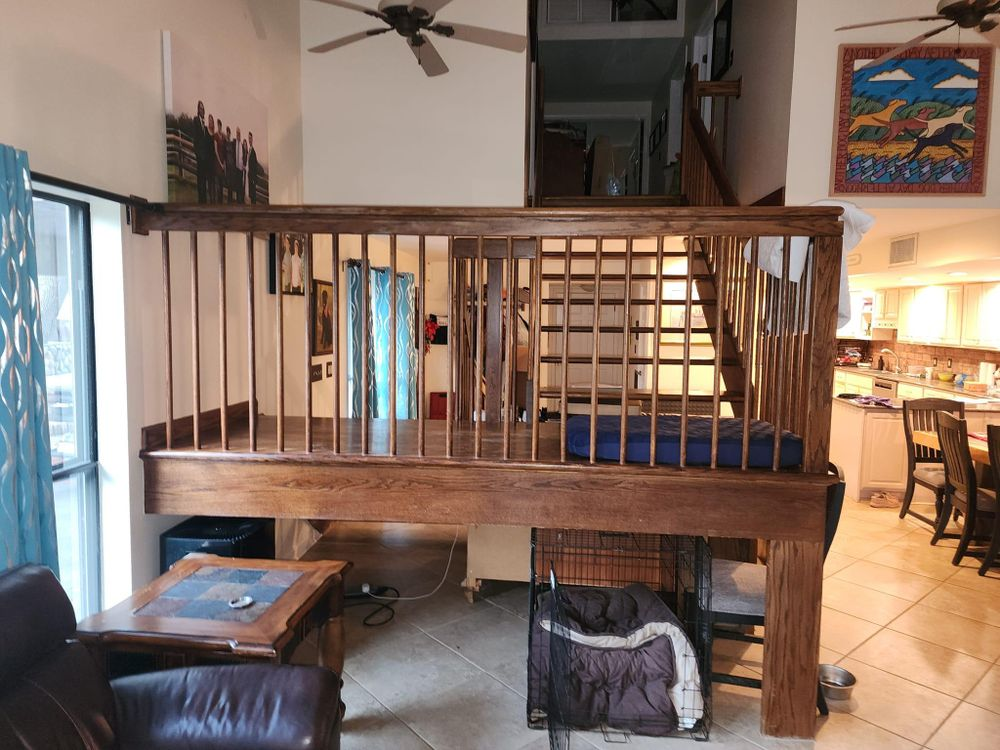 Loft bed with wooden railings, stairs. Living room with a dog crate beneath, kitchen in background.