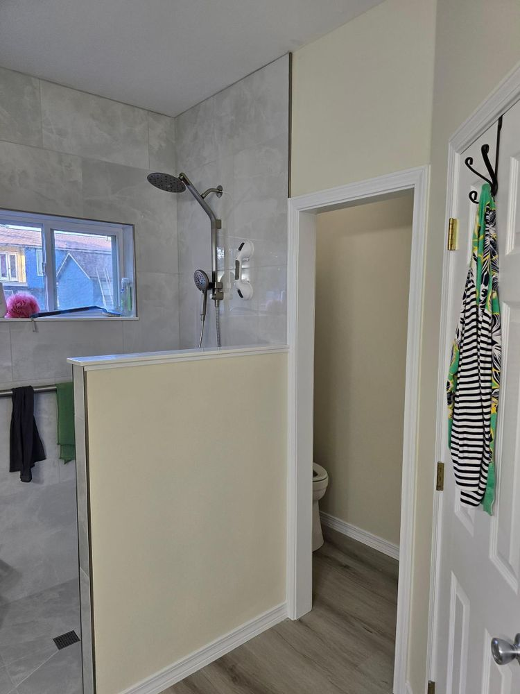 Bathroom with shower, toilet, and doorway. Shower has gray tile, tan wall. Door has clothes hanging.