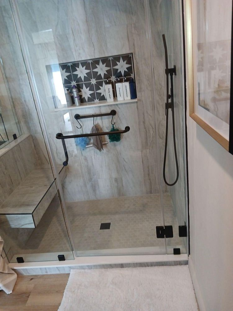 Modern shower with glass door, gray tile walls, and black fixtures; a white rug on the floor.