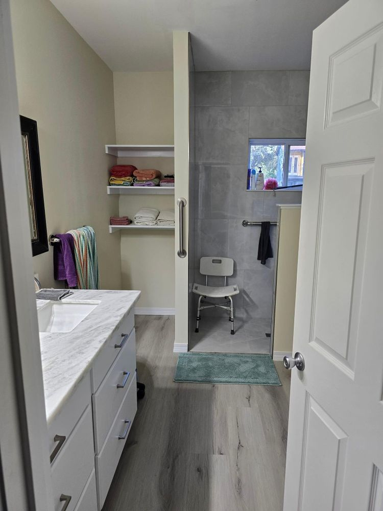 Bathroom with accessible shower, vanity, shelving, and open door. Light wood floors and neutral walls.