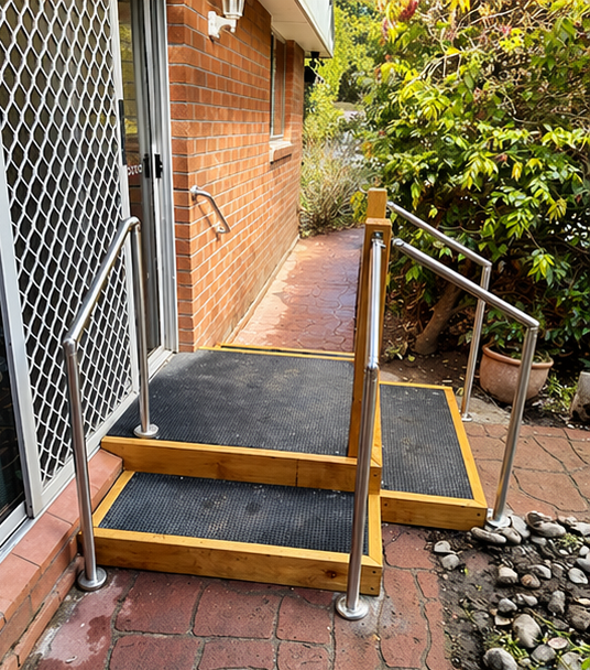 Wooden porch steps with black anti-slip treads and silver handrails beside a brick house walkway
