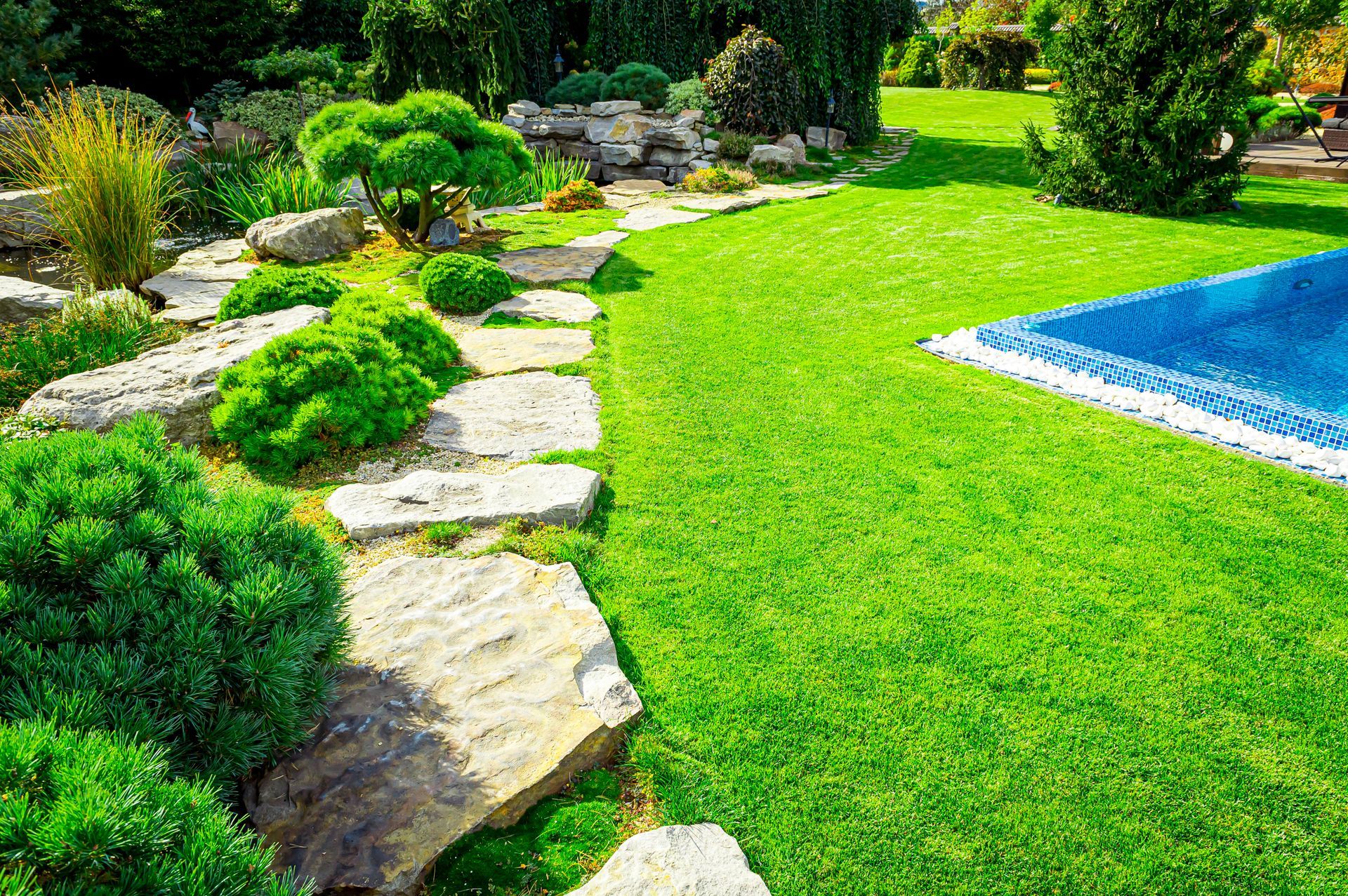 Stone path with lush green grass and bushes, adjacent to a pool.
