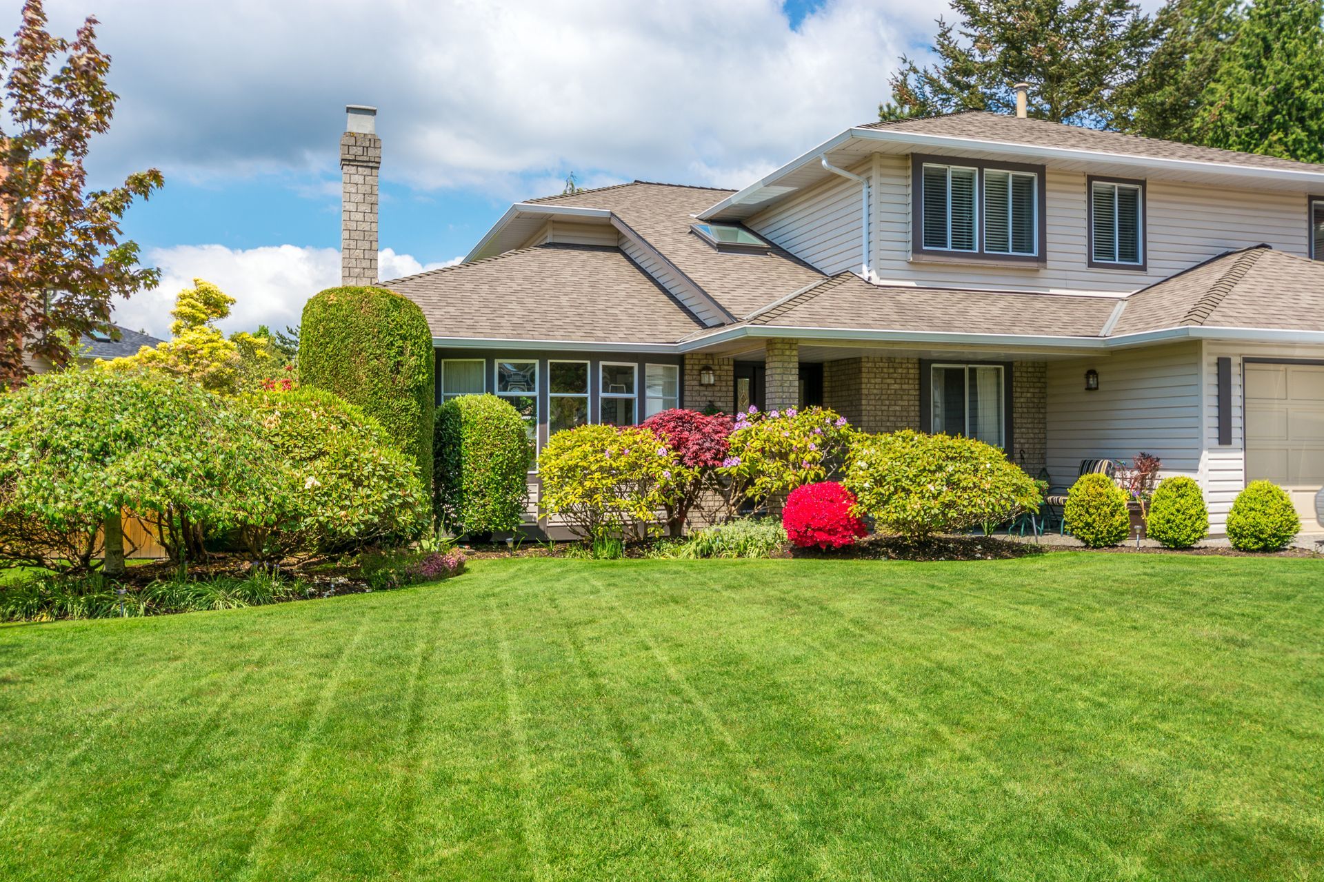 Two-story house with green lawn, various green and red bushes, and a blue sky.