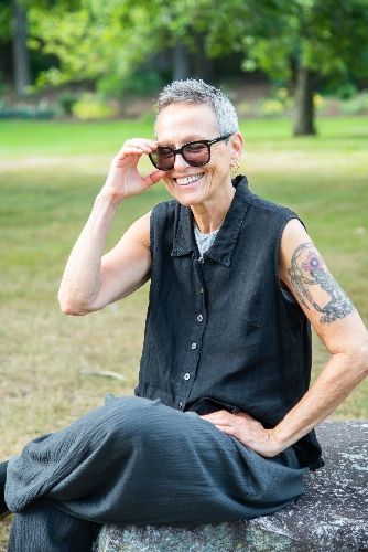 Person sitting on a rock in a park, smiling and adjusting sunglasses, wearing a sleeveless black shirt.