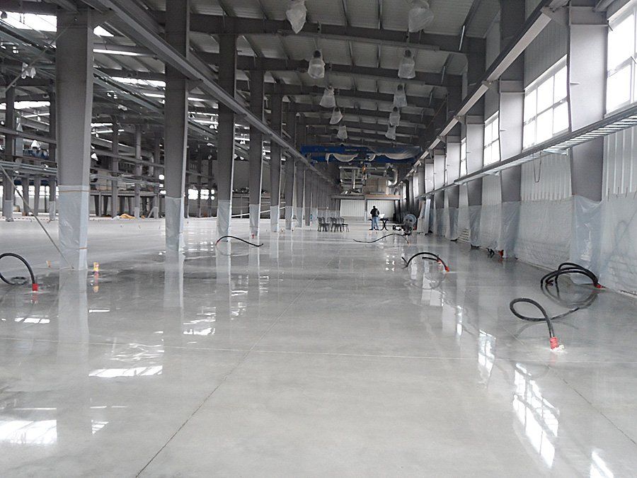 Large industrial interior with polished concrete floor, steel beams, and windows. Workers are present.