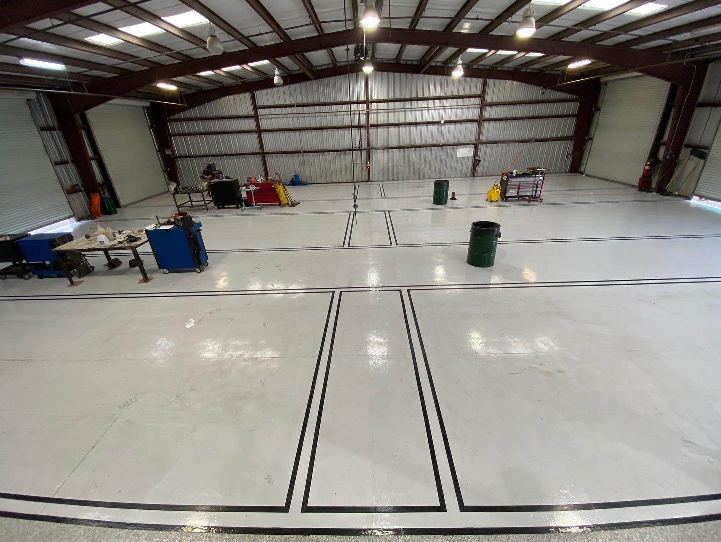 Large indoor space with gray metal frame, light-colored floor, and black taped lines. Equipment and trash cans scattered.