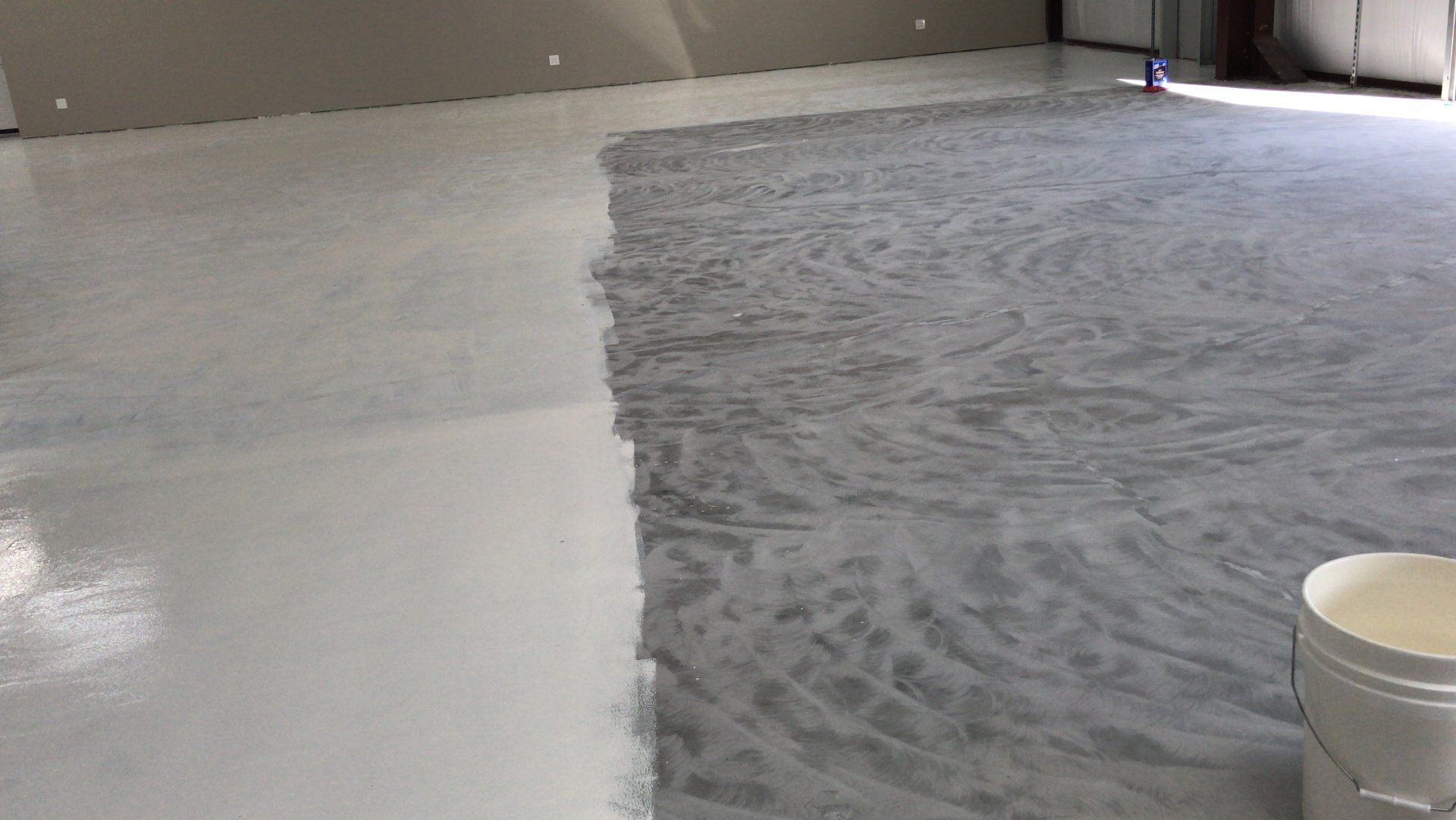 A floor being coated with light gray epoxy. Left side is finished, right side is in progress, with a bucket visible.