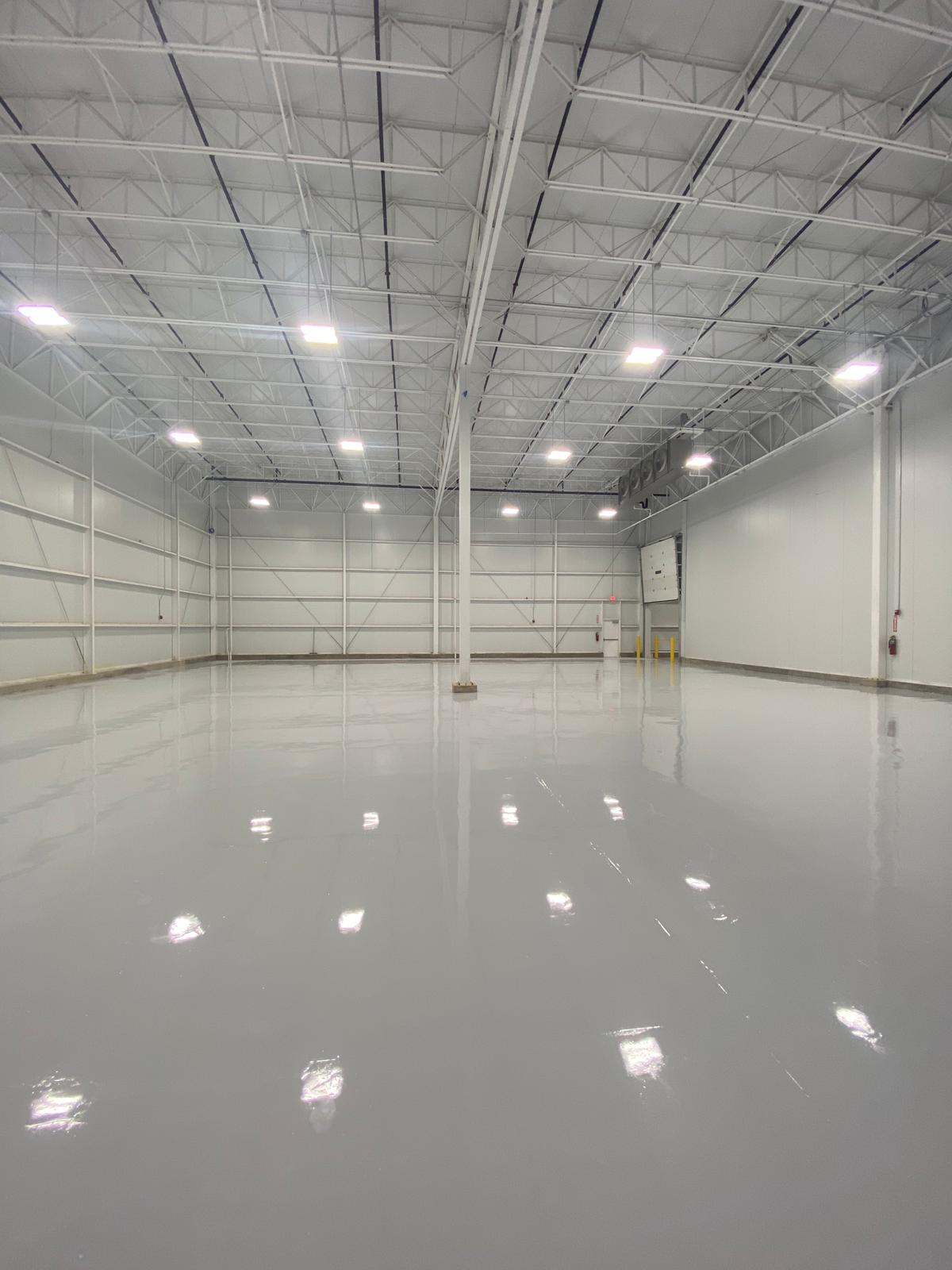 Empty, spacious warehouse interior with a gray, polished floor, bright lights, and white walls.