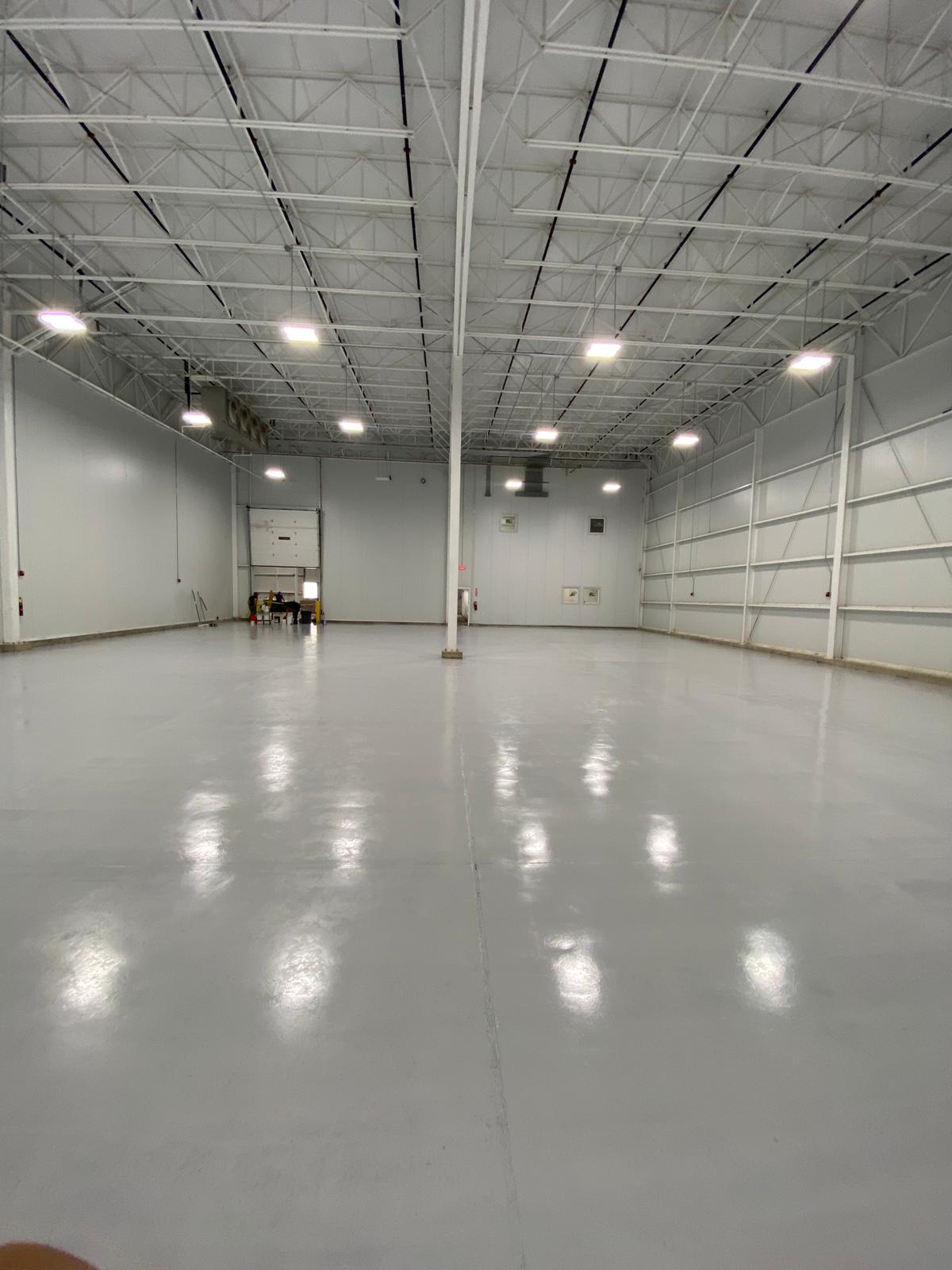 Empty, gray-floored warehouse interior with overhead lighting, metal beams, and a loading door in the distance.