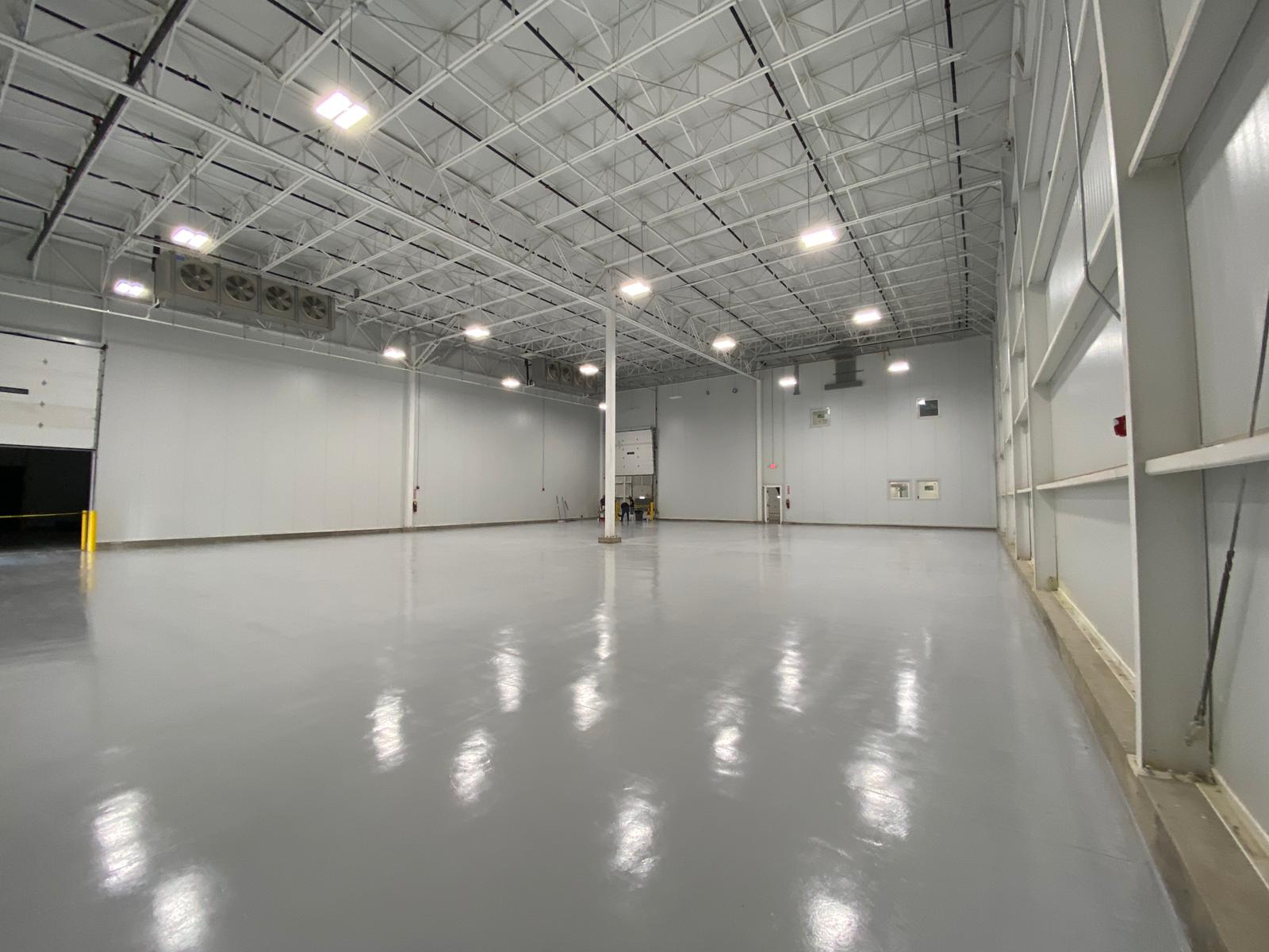 Large, empty warehouse interior; gray walls and floor, white ceiling with lights, metal beams.
