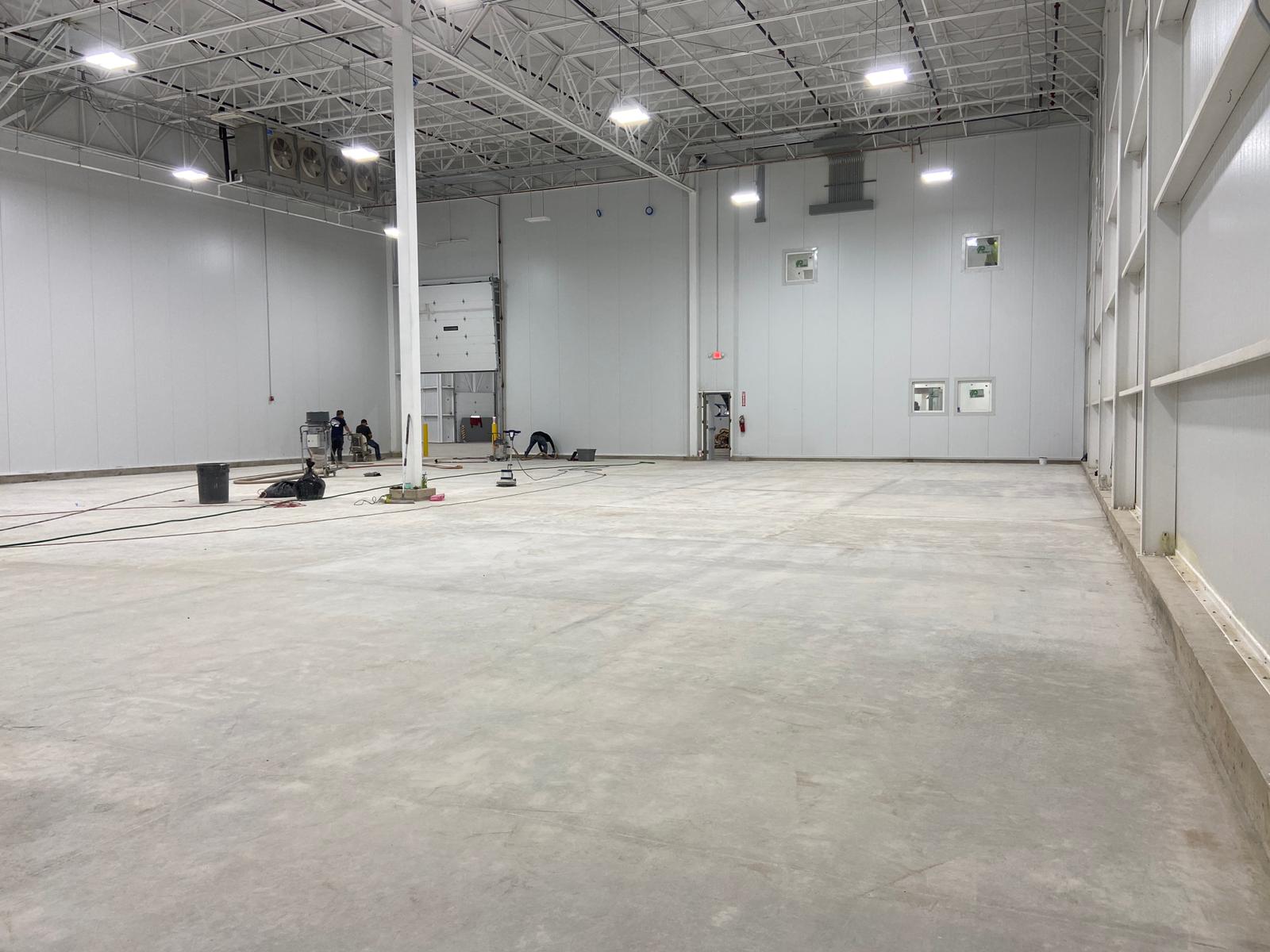 Large, empty industrial warehouse with white walls, concrete floor, and few workers.