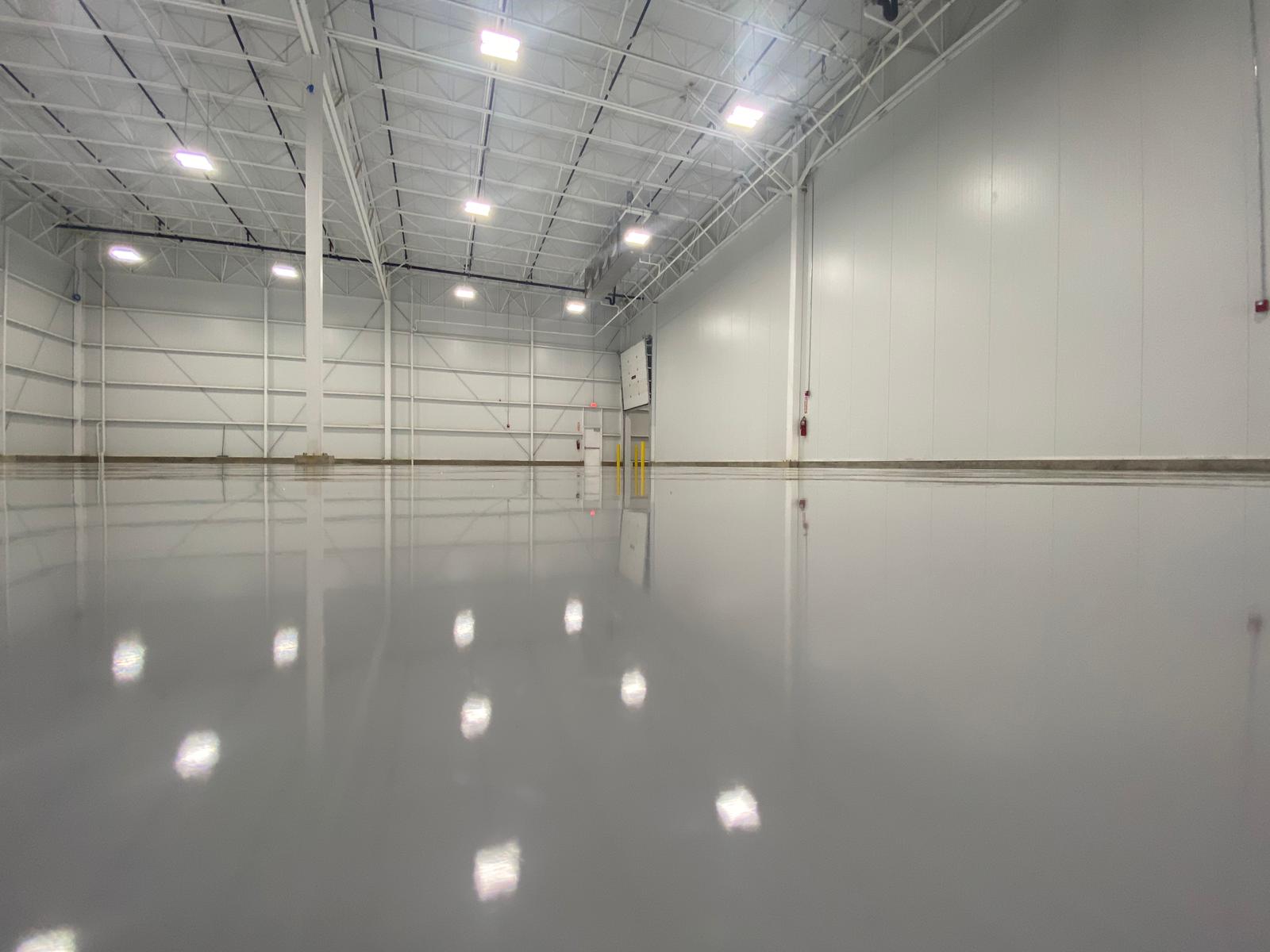 Shiny gray concrete floor in a large, empty warehouse with white walls, ceiling, and overhead lighting.