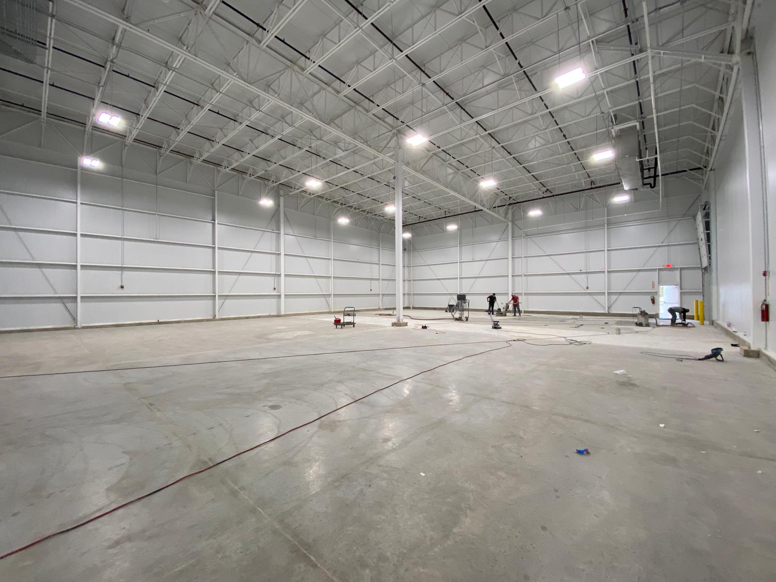 Large, empty industrial warehouse with high ceilings, white walls, and bright overhead lights. Concrete floor.