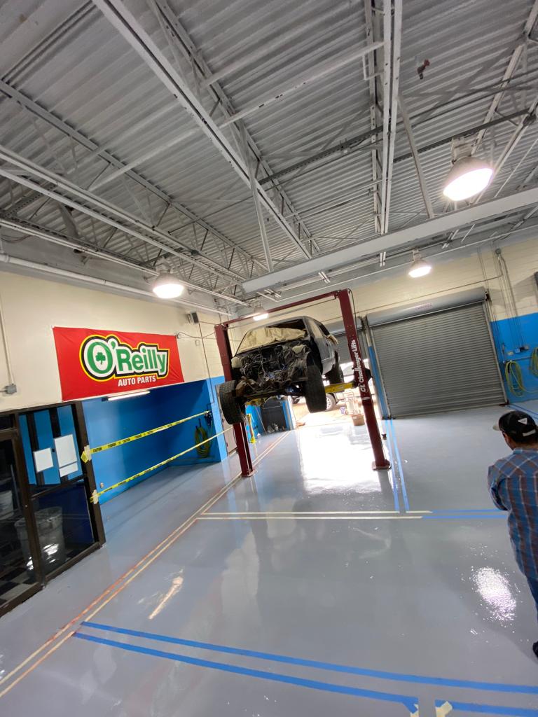 Car on a lift in a repair shop. Red lift, white ceiling, blue/gray floor. Person on the right.
