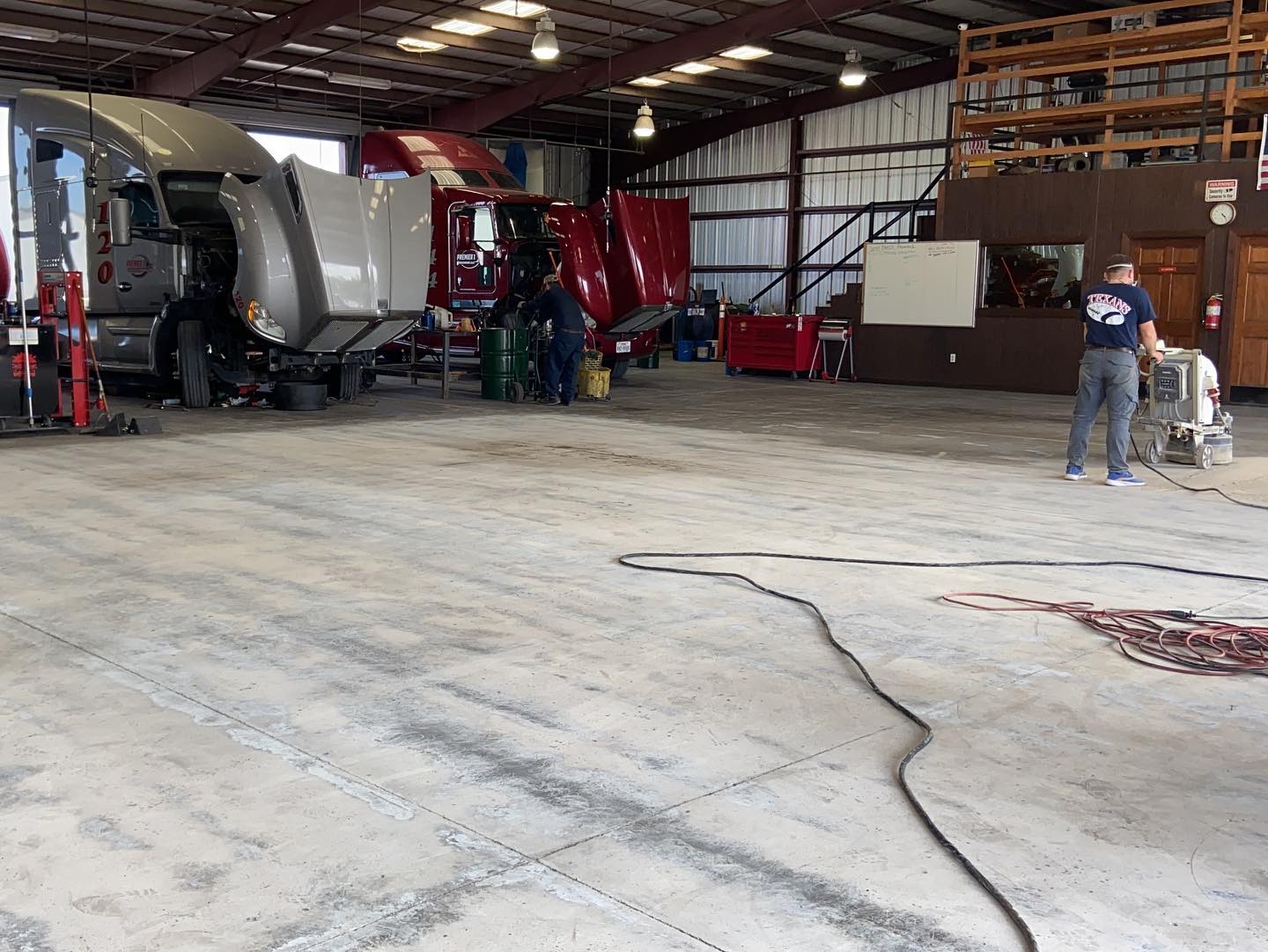 Mechanics working on trucks inside a large garage. One person uses a floor polisher.
