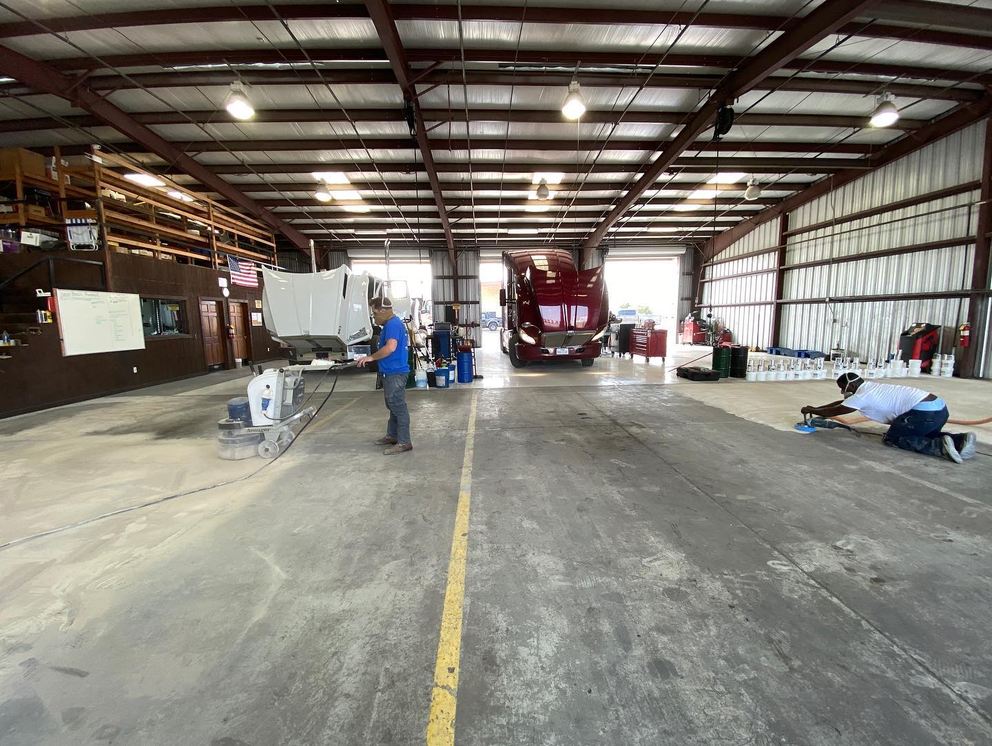 Inside a truck repair shop, a red semi-truck is centered, two workers perform maintenance.