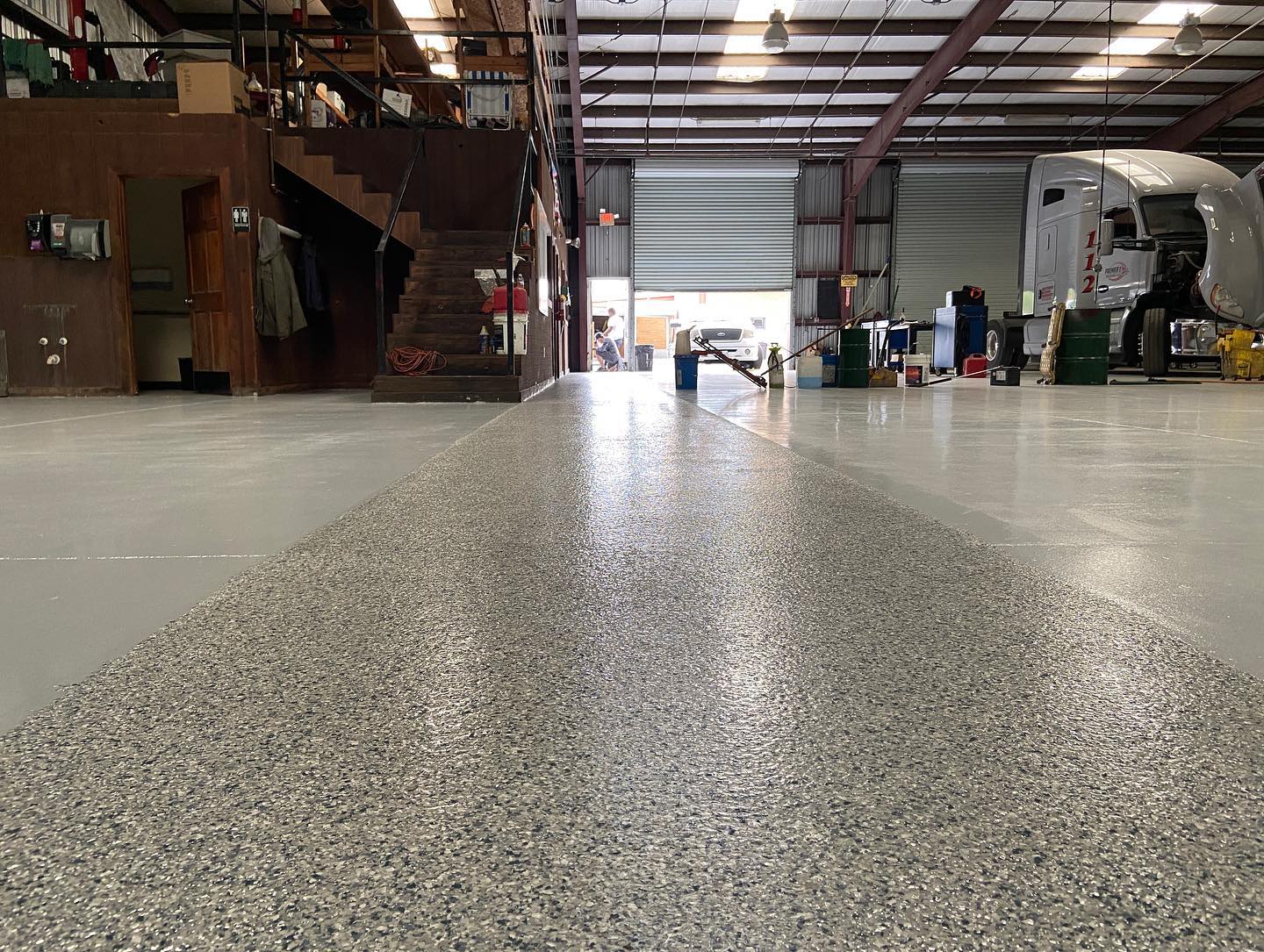 Interior of a warehouse with a newly coated epoxy floor. The open door shows a truck. A staircase is visible.