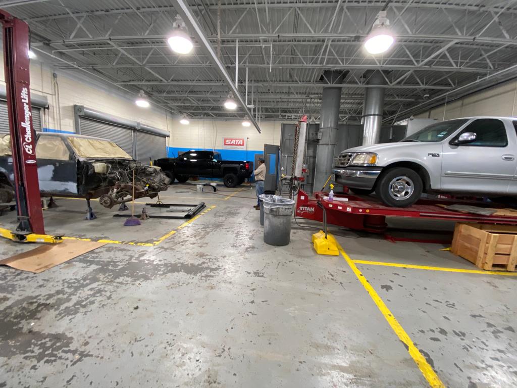 Auto repair shop interior with vehicles in various states of repair. A lifted silver truck and a stripped-down chassis are visible.