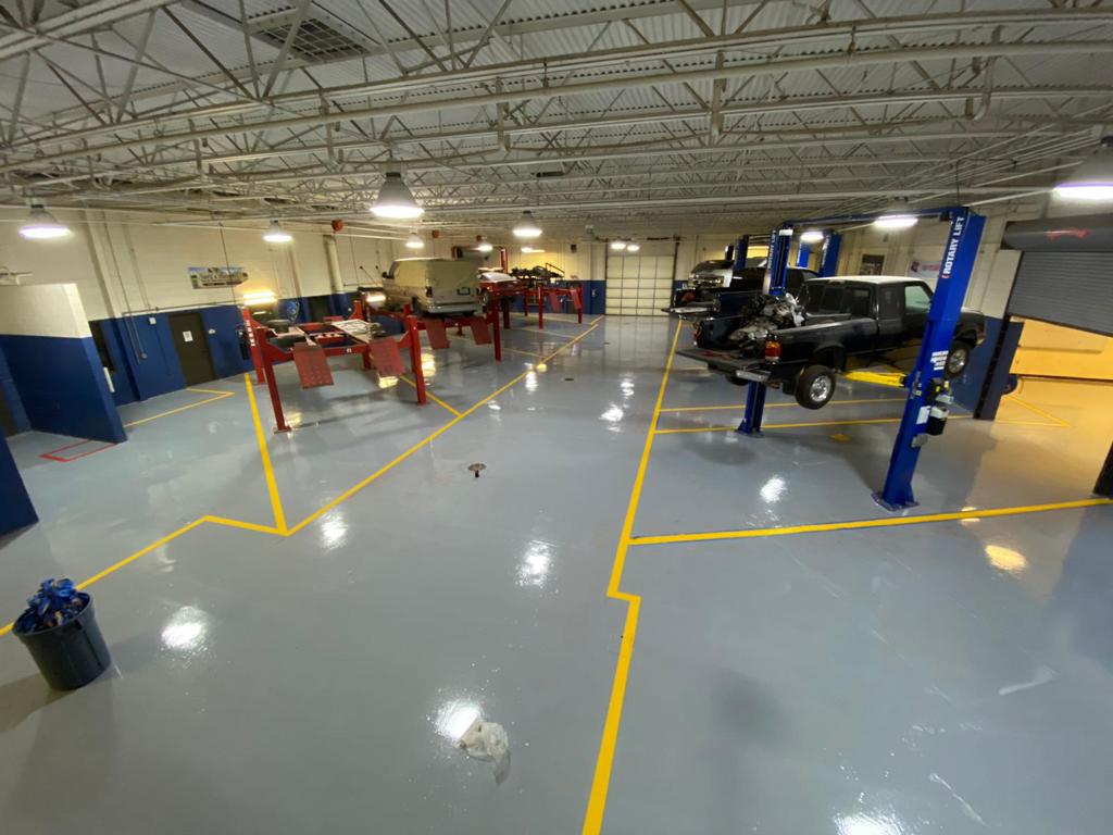 A spacious auto repair shop with multiple vehicles on lifts. Grey floor with yellow markings and overhead lights.