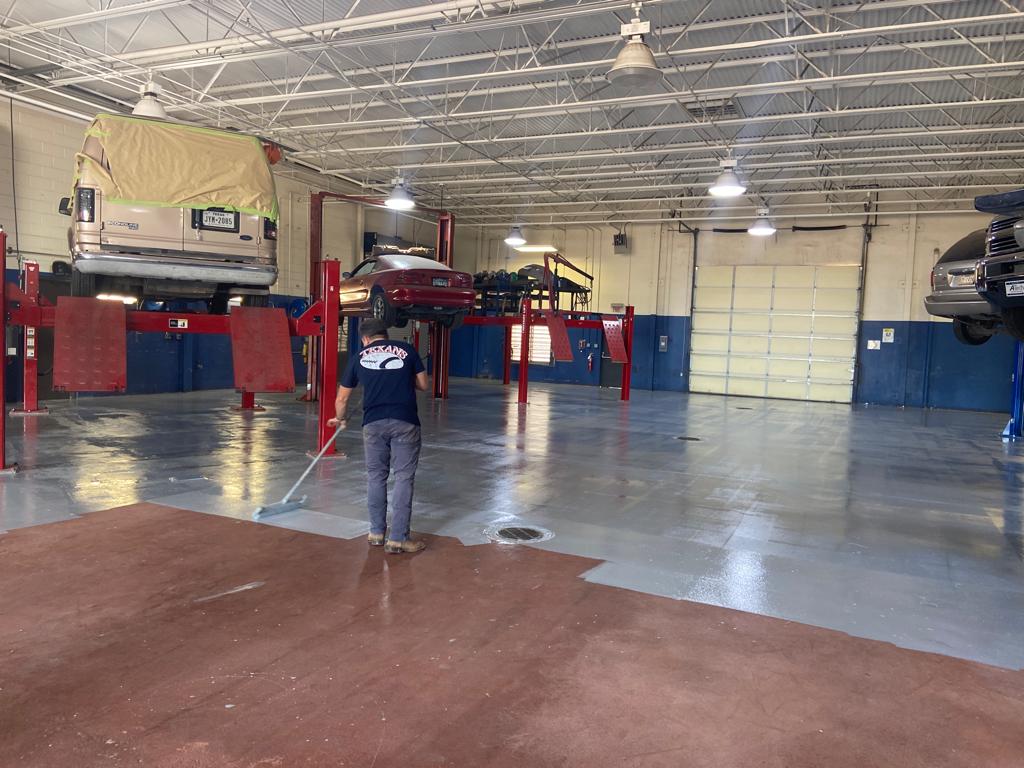 Person applying a red coating to a garage floor. Cars on lifts are in the background.