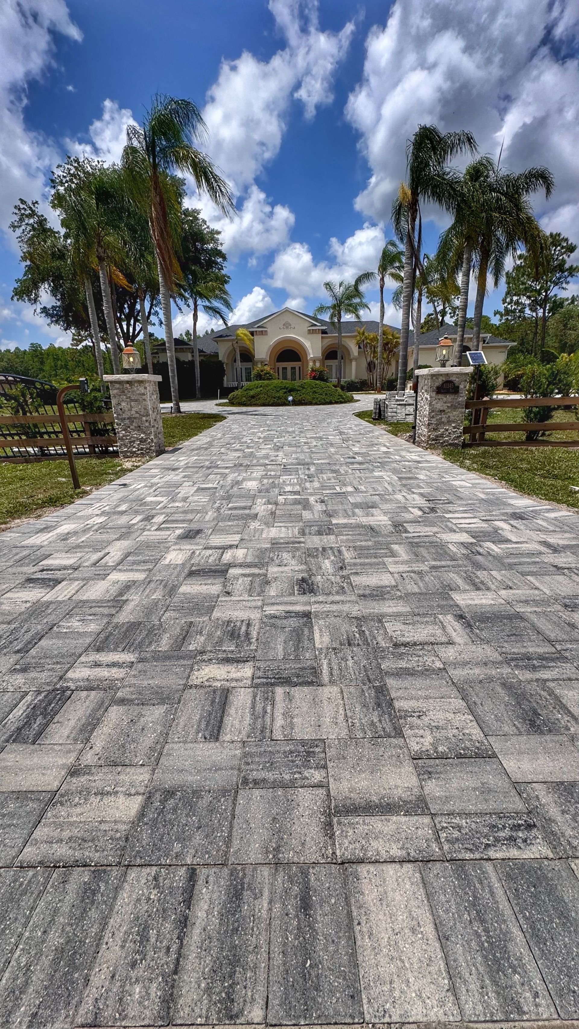 Brick driveway leading to a beige house with palm trees under a blue sky with clouds.