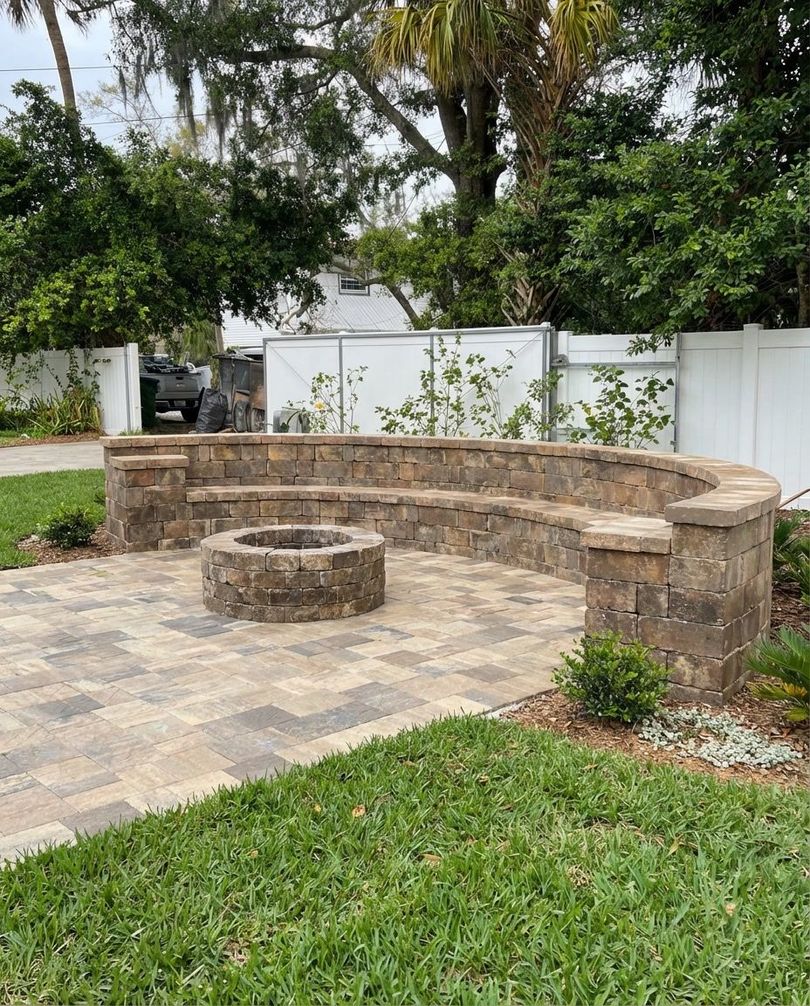 Stone patio with built-in curved seating and a central fire pit, surrounded by green grass and landscaping.