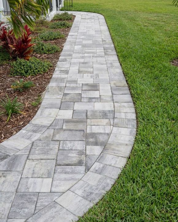 Wide brick paved driveway leading to a single-story gray house with green lawn and palm trees