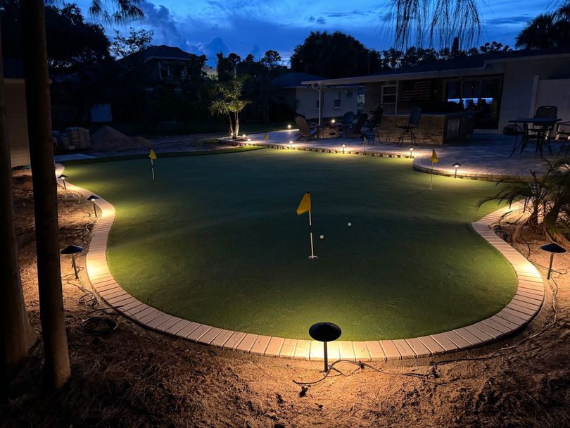 Backyard putting green at night, lit by landscape lights. Yellow flags mark the holes.