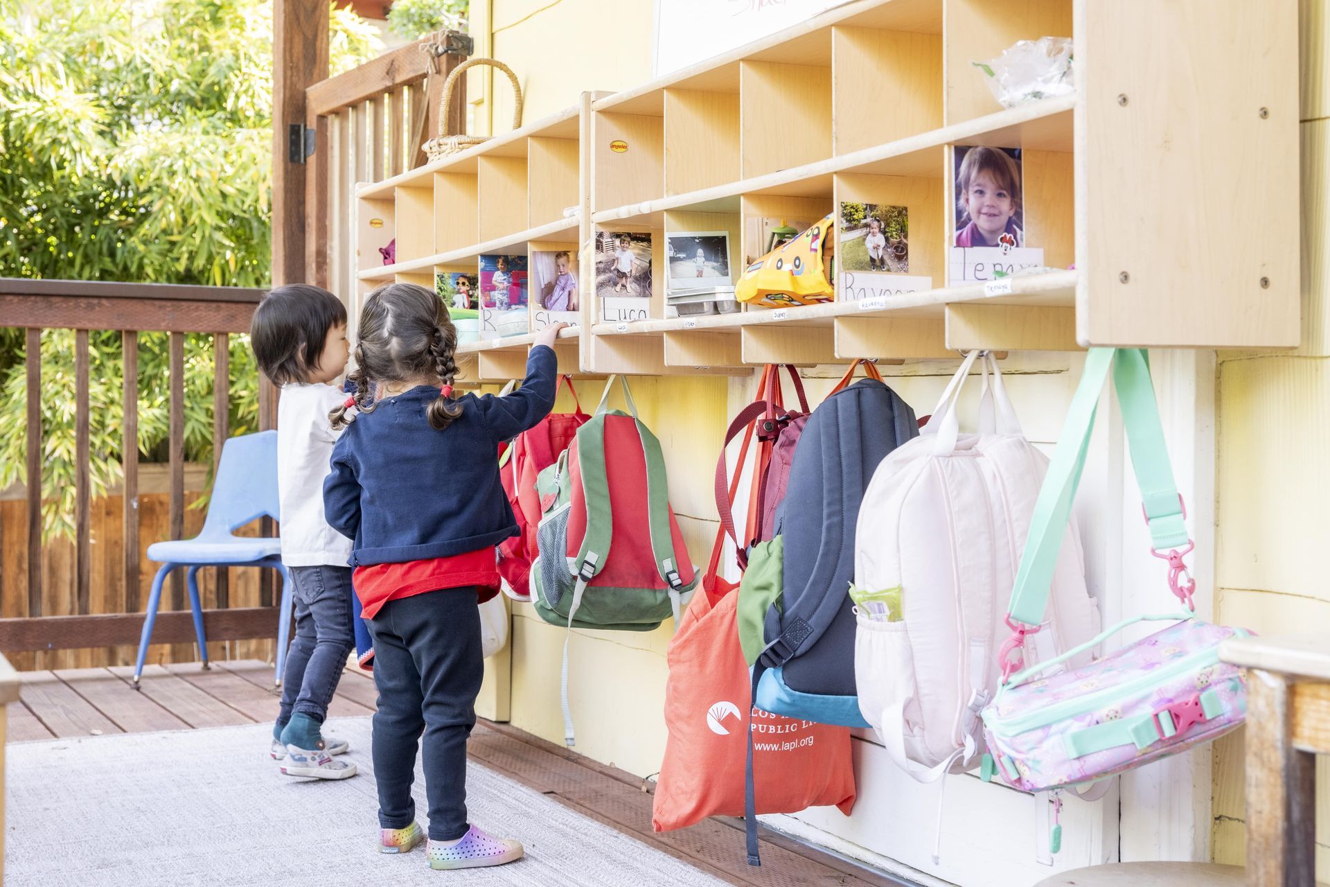 Montessori children are standing in front of a shelf with backpacks hanging on it.